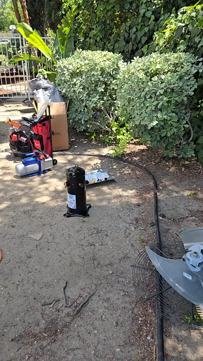 A burned-out air conditioner compressor standing in the dirt of a residential backyard, with the condenser fan blade and guard lying on the ground beside it