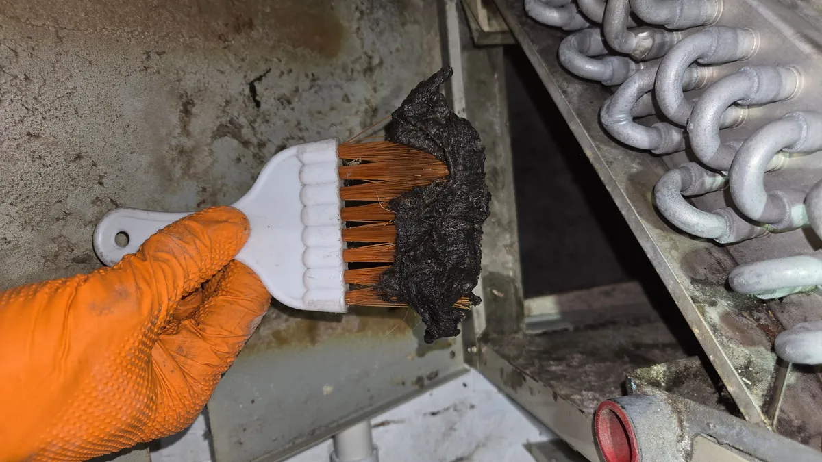Gloved hand holding a brush caked with a thick black mat of dirt pulled off an evaporator coil, with the coil's tube bends behind it