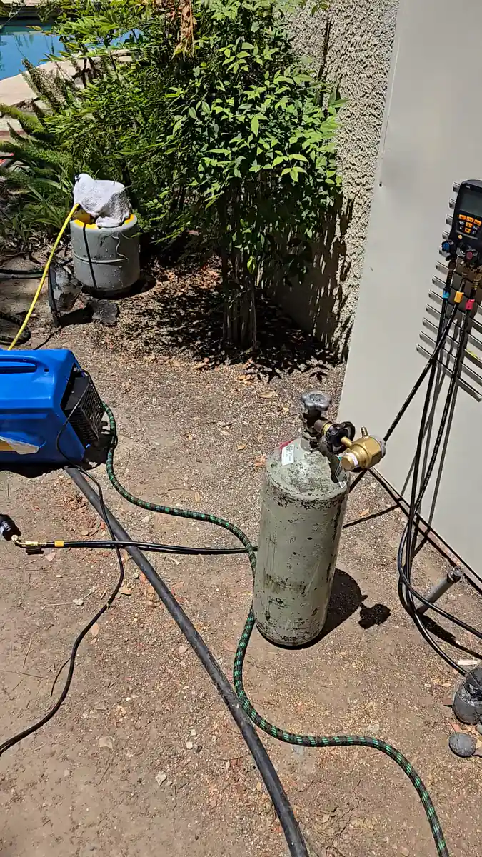 A nitrogen cylinder with its regulator, digital manifold gauges hanging on the wall and a refrigerant cylinder on a yellow hose set up beside the unit