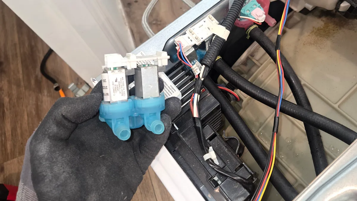 Two-coil water inlet valve held in a gloved hand over an opened Maytag washer