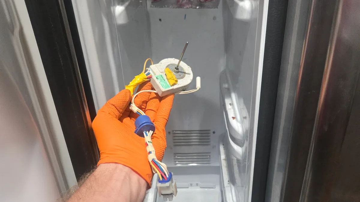 Evaporator fan motor with its wiring harness held up in a gloved hand inside the emptied freezer of a Whirlpool side-by-side