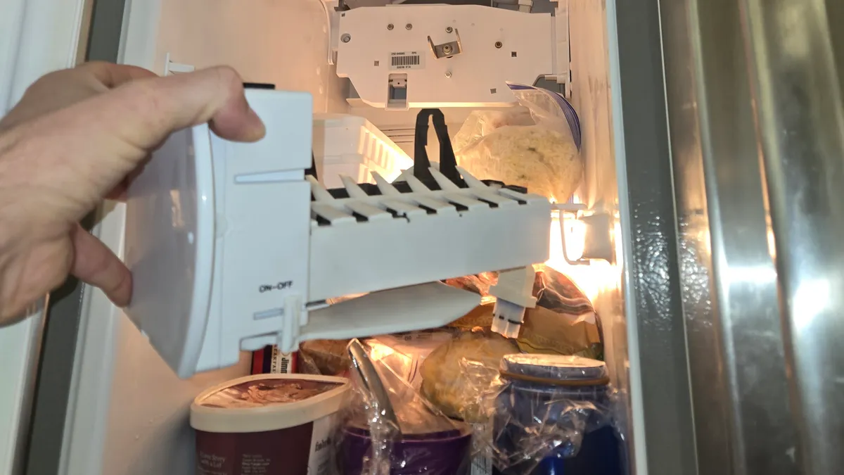 Ice maker held in front of the open freezer of a Whirlpool side-by-side refrigerator, with food still on the shelves