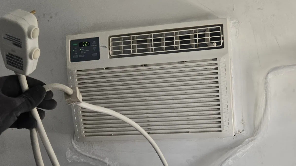 Gloved hand holding the safety plug of a window air conditioner's cord, with the unit set in the wall behind it