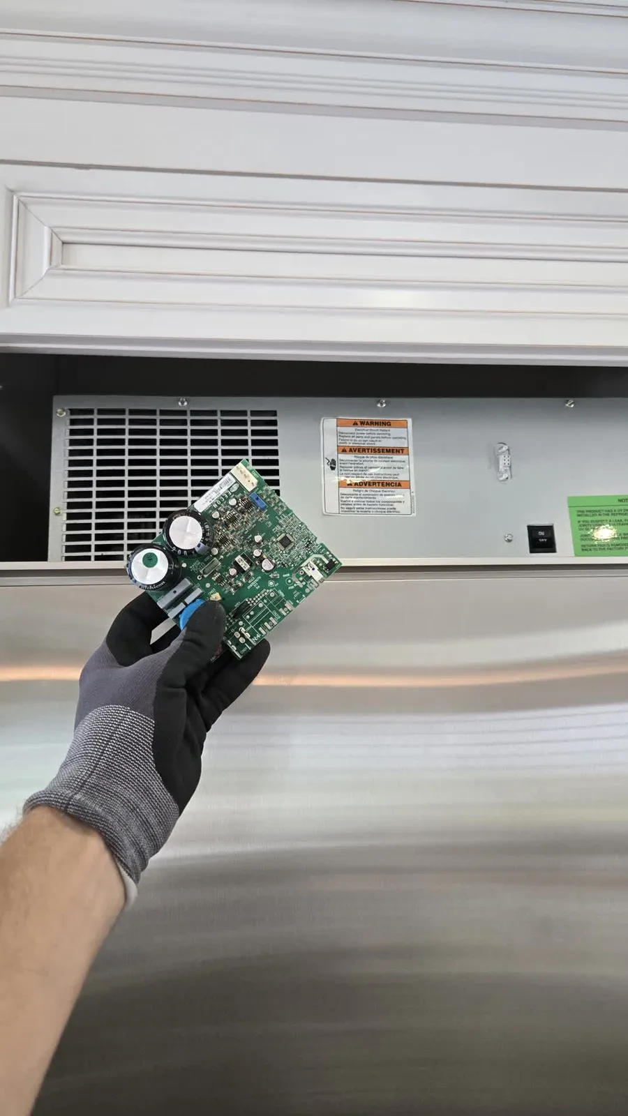New Embraco inverter board held in a gloved hand in front of the top grille of a built-in KitchenAid refrigerator
