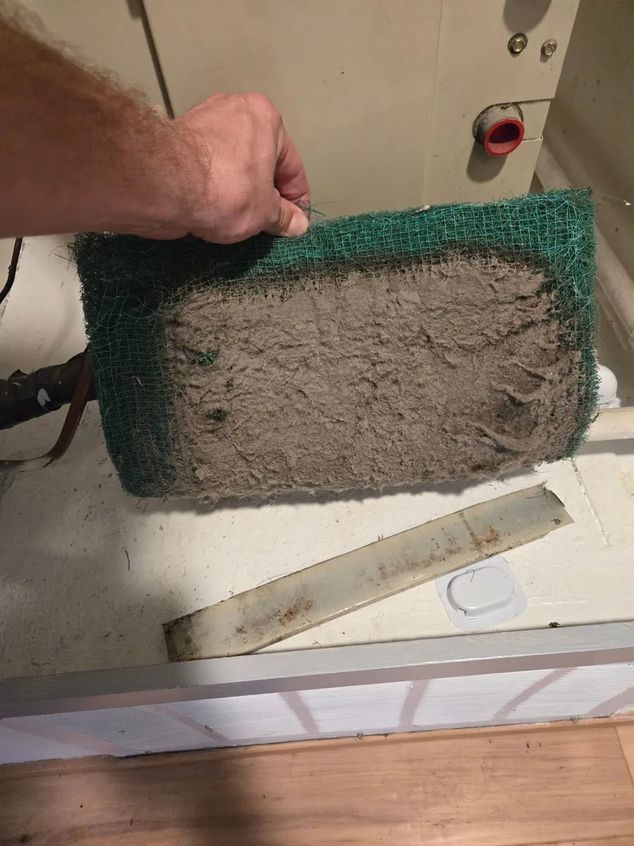 Hand holding a green mesh filter pulled from an air handler, its face matted with a thick layer of gray dust