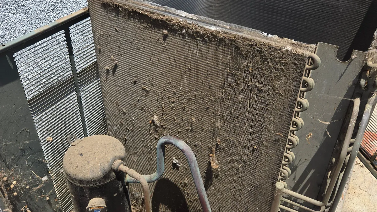 AC condenser with its top removed before cleaning, the coil coated in a gray layer of dust and debris and dirt collected around the compressor