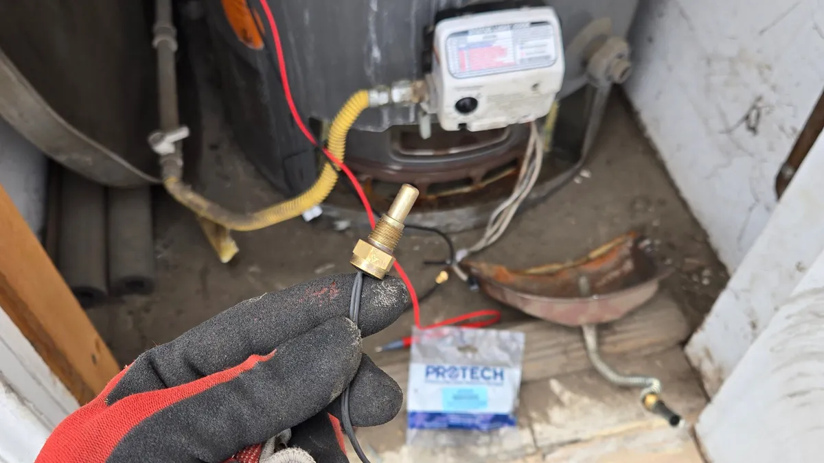 Gloved hand holding a small brass chamber sensor on a thin black lead in front of a gas water heater's control valve, with a ProTech parts bag on the floor