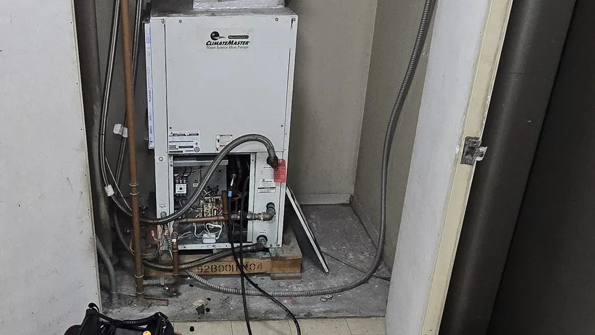 ClimateMaster water-source heat pump in a building utility closet, its lower service panel off and a refrigerant gauge hose connected