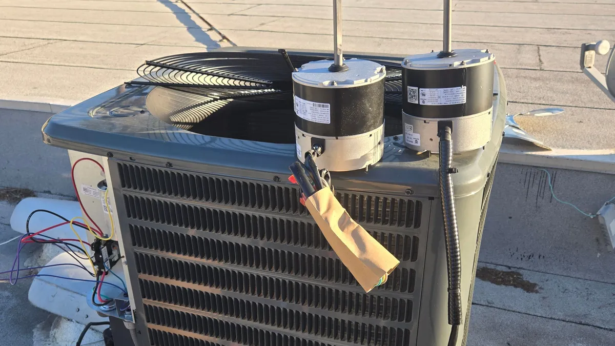 Two condenser fan motors resting on top of a Goodman condenser on a flat roof, with the fan guard lifted off during the motor replacement