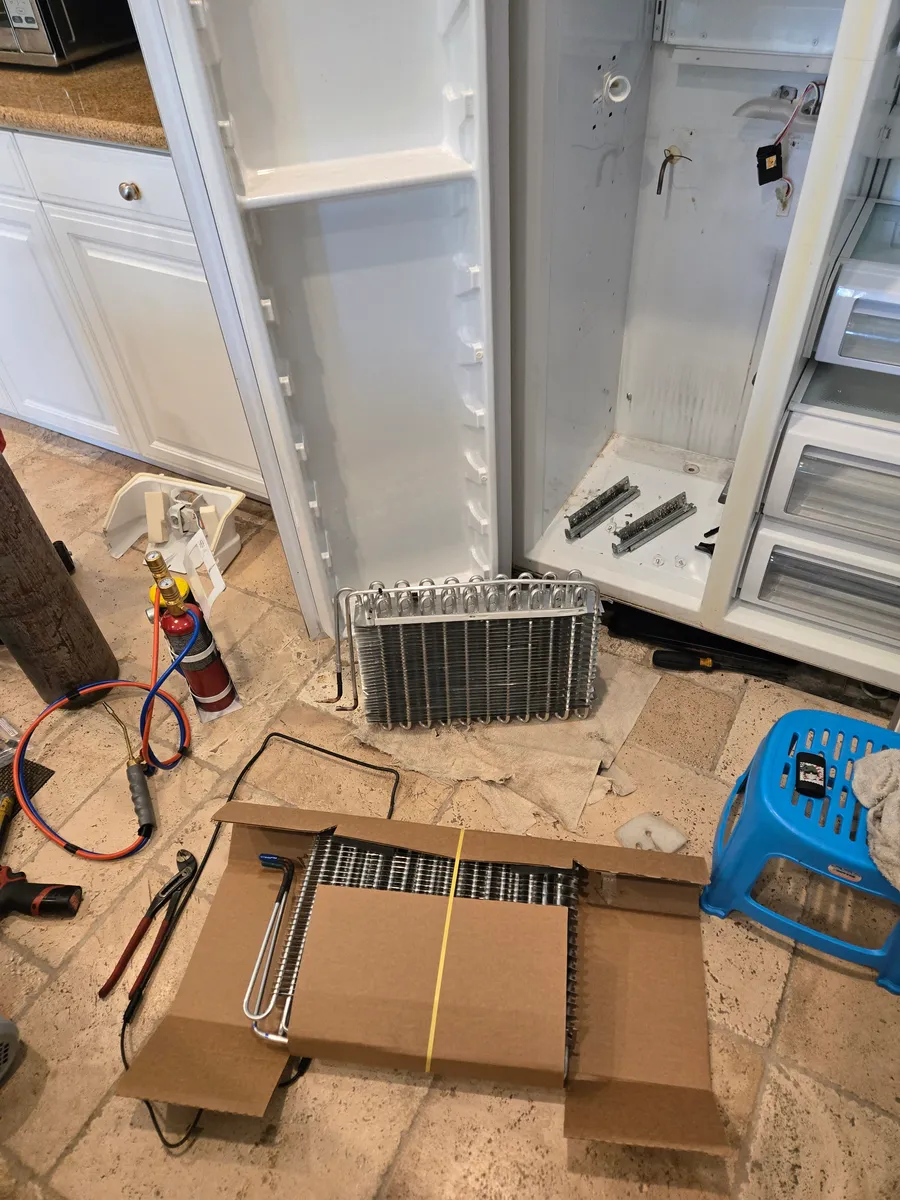 The removed evaporator coil on a towel and the new one still in its box, in front of an opened built-in KitchenAid refrigerator