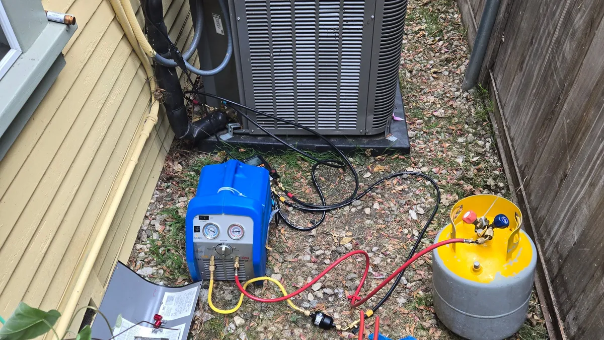 A Lennox outdoor unit with its corner panel off and a refrigerant recovery machine and recovery cylinder hooked up with hoses during a compressor replacement