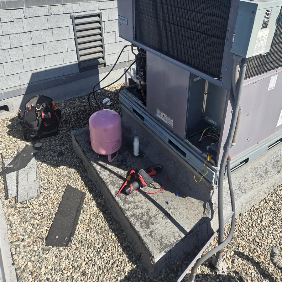 Packaged AC unit on a rooftop pad with its side panels off, hoses connected and a refrigerant cylinder beside it