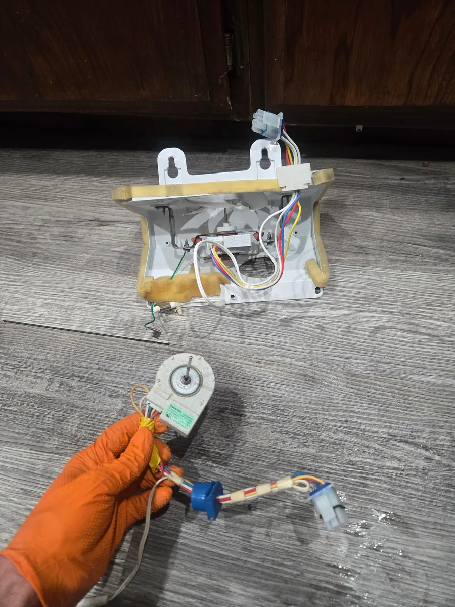 Whirlpool refrigerator evaporator fan housing on the floor, and the fan motor with its wiring harness in a gloved hand